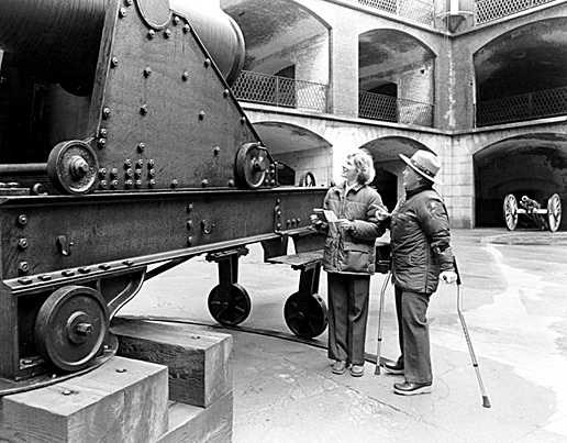 A ranger using forearm crutches talks with a visitor beside a large cannon.