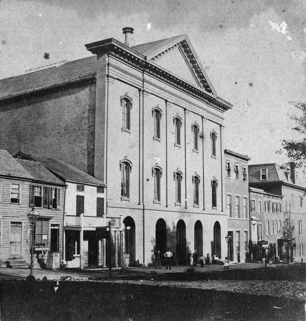 A three story building with an imposing facade in a city block of smaller buildings.