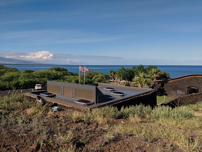 The repaired comfort station, with a flat roof and skylights, among dry grass and new plantings, the ocean beyond