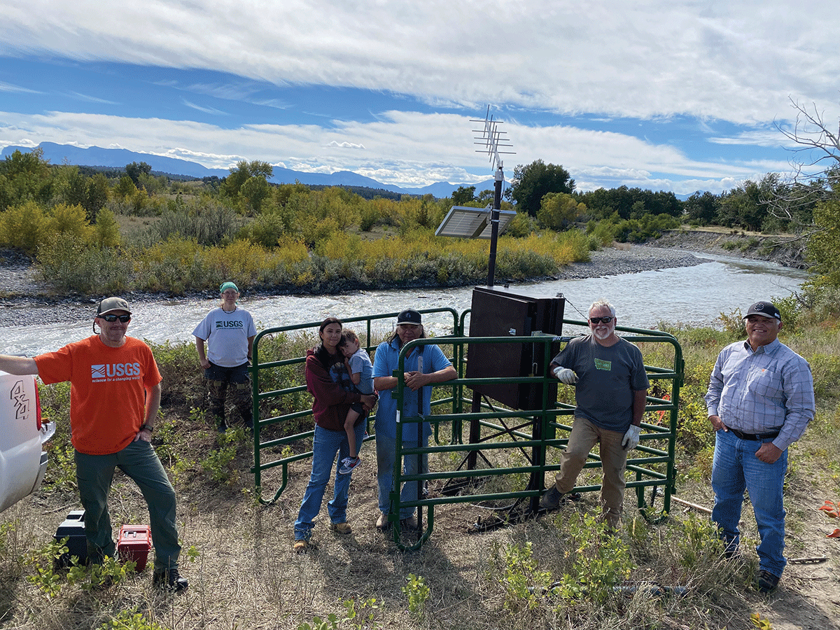 Six adults and a child standing by a streamgage with a river behind them.