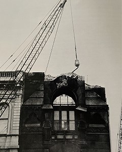 A tall building being torn down, with a crane and wrecking ball.