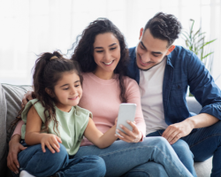 a mother, father, and daughter sitting close on a couch while looking at a cellphone
