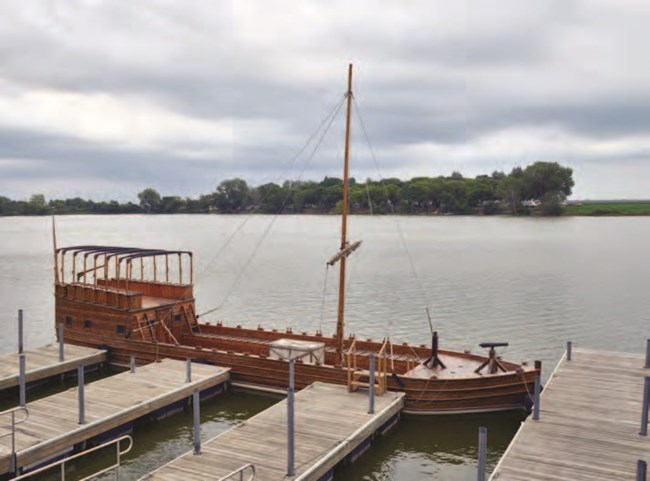 A long wooden keelboat with a small rear cabin and a single mast, tied up at a dock on a calm river