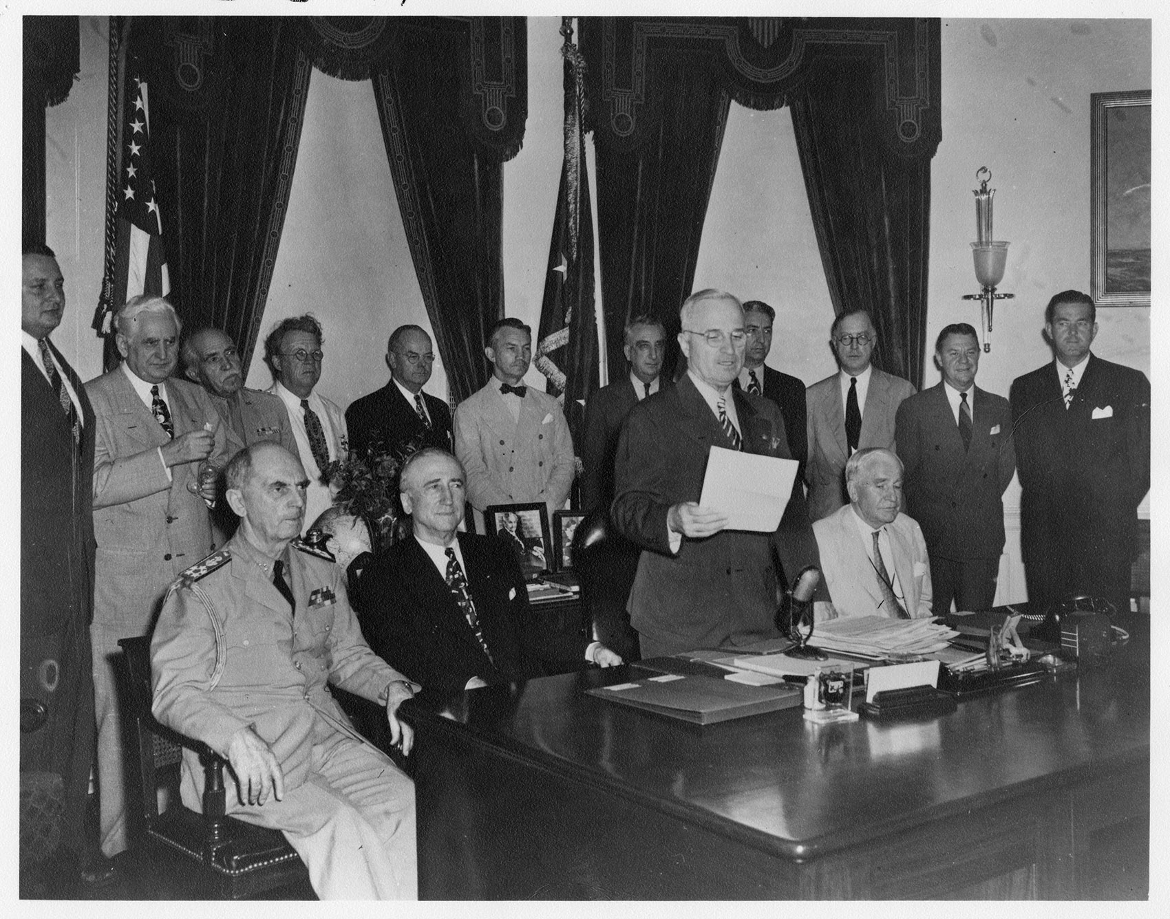 President Truman at his desk reading a statement, surrounded by officials and reporters