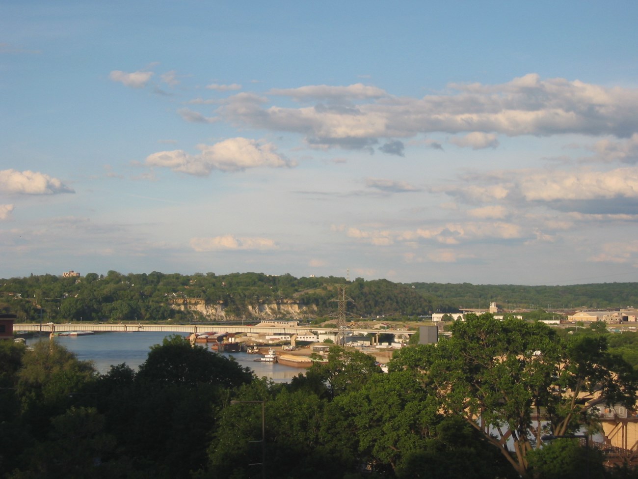 Buildings, pavement and a bridge along and over the Mississippi River, with green trees in the foreground and on the horizon beyond the road bridge.