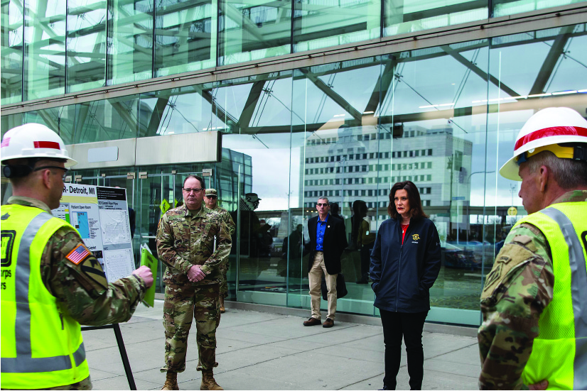 A photo shows Gretchen Whitmer standing outside of an urban building being briefed by a person in military uniform. Two other people in military uniforms and a plainclothed person are at the briefing.