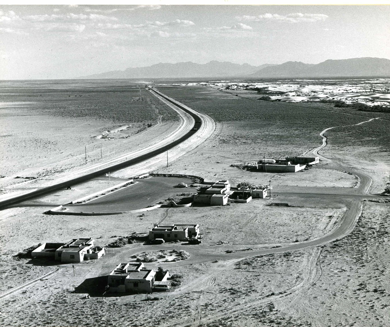 Historic aerial view of White Sands' housing, visitor center and maintenance buildings clustered by a curving road, with the dunes and mountains beyond
