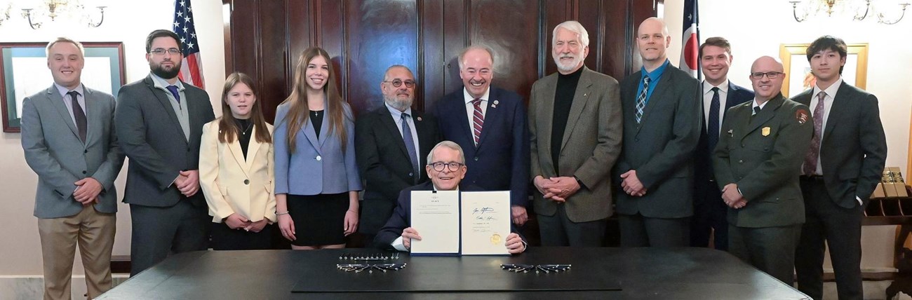 Ohio's governor seated at a table with the signed bill, surrounded by Garfield descendants and officials.