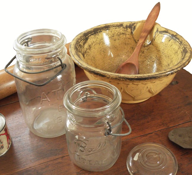 Glass canning jars, a yellow bowl and a wooden spoon on a wooden kitchen table.
