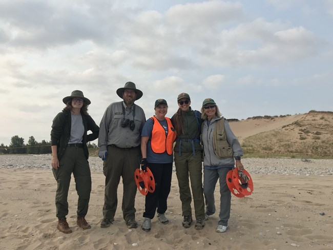 Five people on a beach: three NPS employees in uniform and two volunteers, Mary at far right in a green volunteer cap