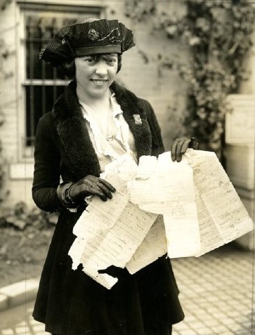 A suffragist holding up the torn, tattered pages of a lobbying list and wearing a small pin.